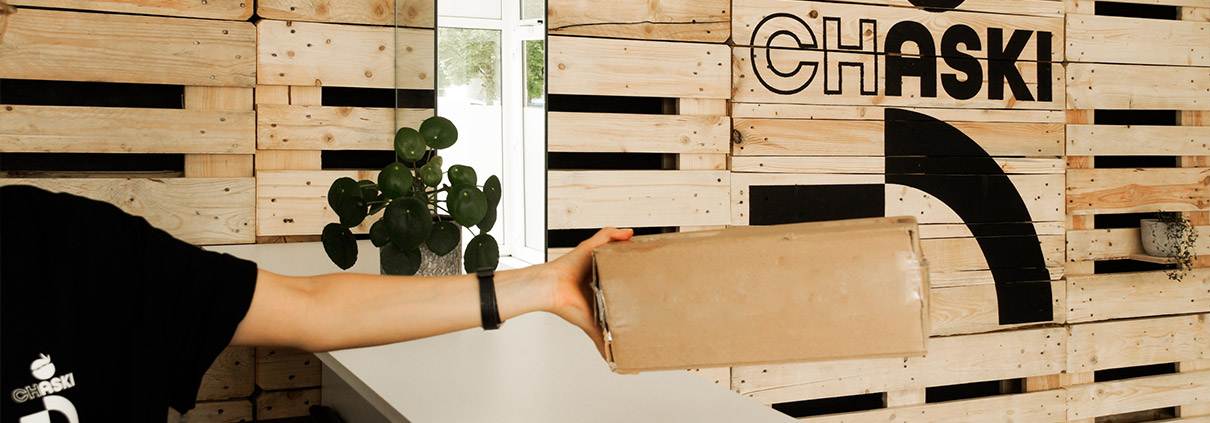 Palettenwand mit Logo con CHASKI, davor der Arm eines Mitarbeiters mit einem Paket in der Hand.