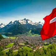 Zollfreigrenze Schweiz 2025: Zollfrei einfuehren Landschaftsbild von einer Stadt im Tal und Berge und ein See im Hintergrund. Im Vordergrund eine schweizer Flagge. Dieses Bild wurde mit KI generiert.