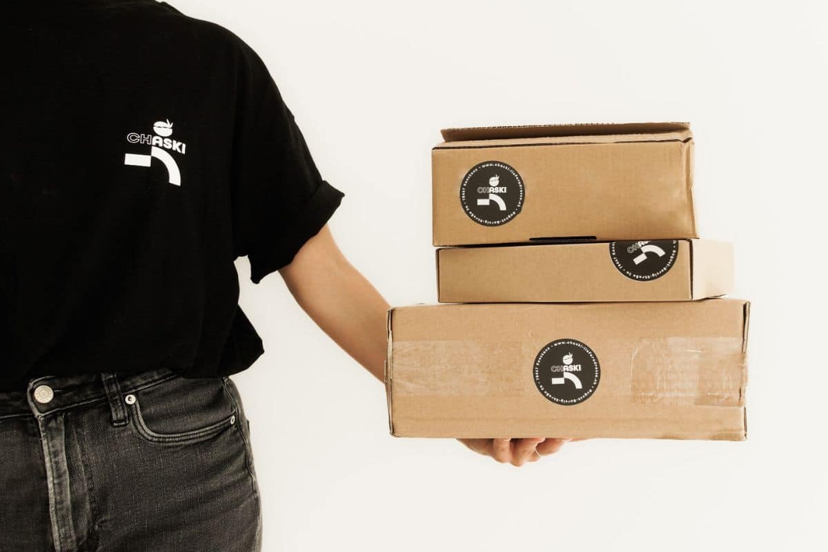 So adressierst du deine Sendung Angeschnittener Mitarbeiter mit CHASKI T-Shirt hält Pakete mit CHASKI Aufklebern in der rechten Hand